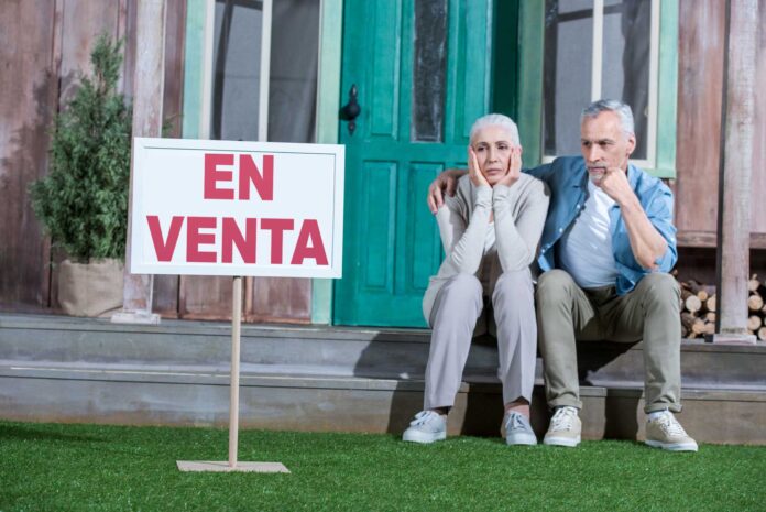 Fotografía de una pareja sentada a la puerta de su casa, con gesto de desánimo al no conseguir venderla.